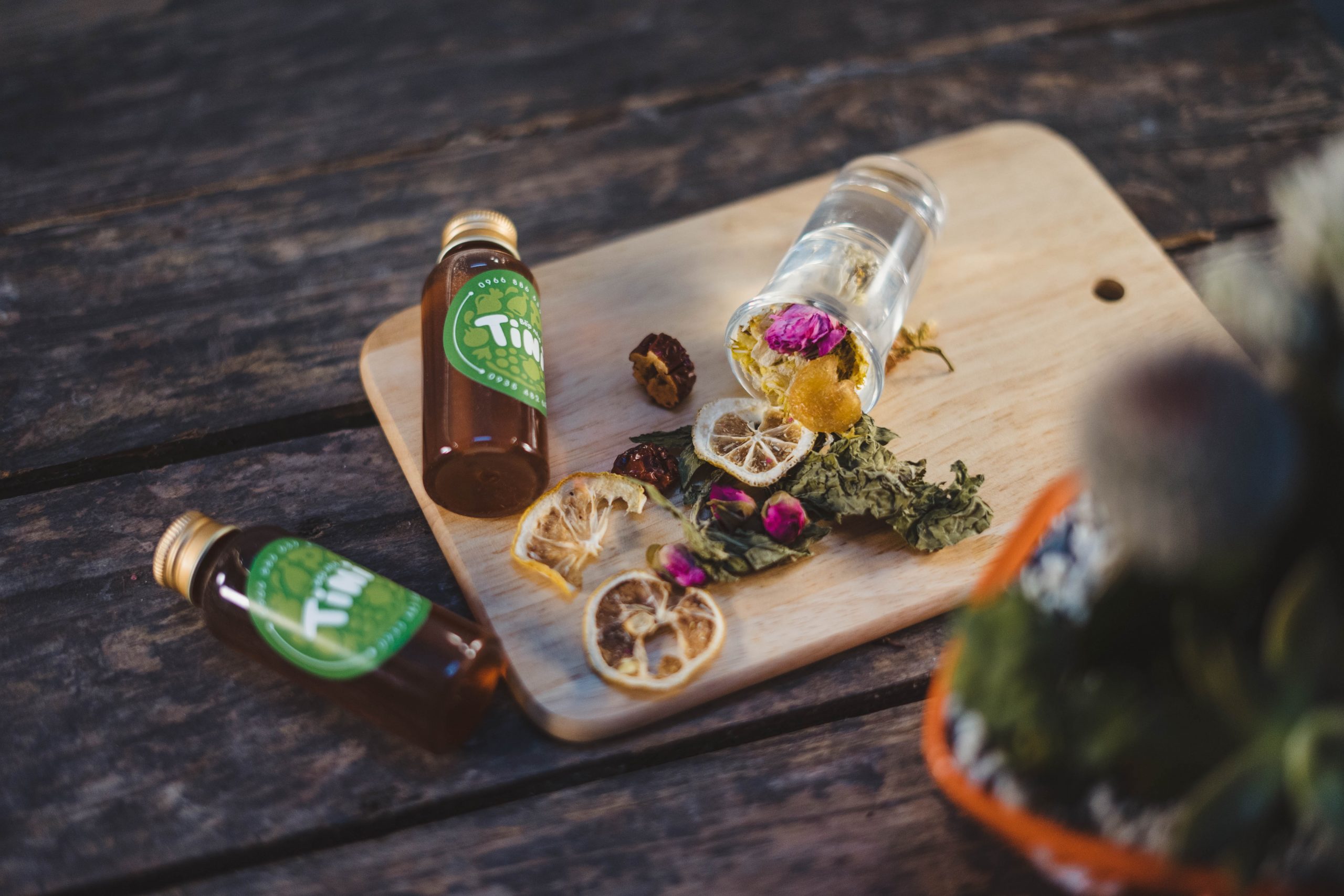 A brown chopping board with orange peels, flowers, and spices, 2 bottles, and a potted plant resting on wood.