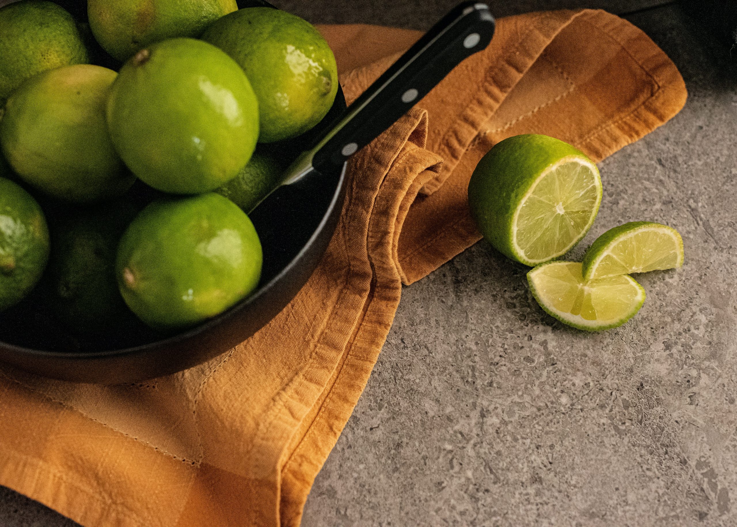 A black pan containing lemons and a knife placed on a yellowish brown cloth with cut pieces of lemon on the side