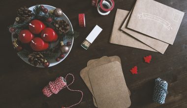 A brown table decorated with two rolls of thread, brown envelops, brown pads, designer tape, two small red Christmas trees, an open matchbox, and a plate with four red candles, pine cones, berries, and baubles.