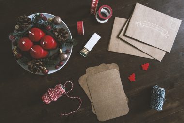 A brown table decorated with two rolls of thread, brown envelops, brown pads, designer tape, two small red Christmas trees, an open matchbox, and a plate with four red candles, pine cones, berries, and baubles.