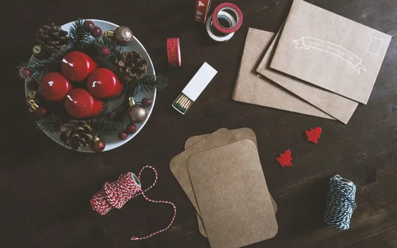 A brown table decorated with two rolls of thread, brown envelops, brown pads, designer tape, two small red Christmas trees, an open matchbox, and a plate with four red candles, pine cones, berries, and baubles.