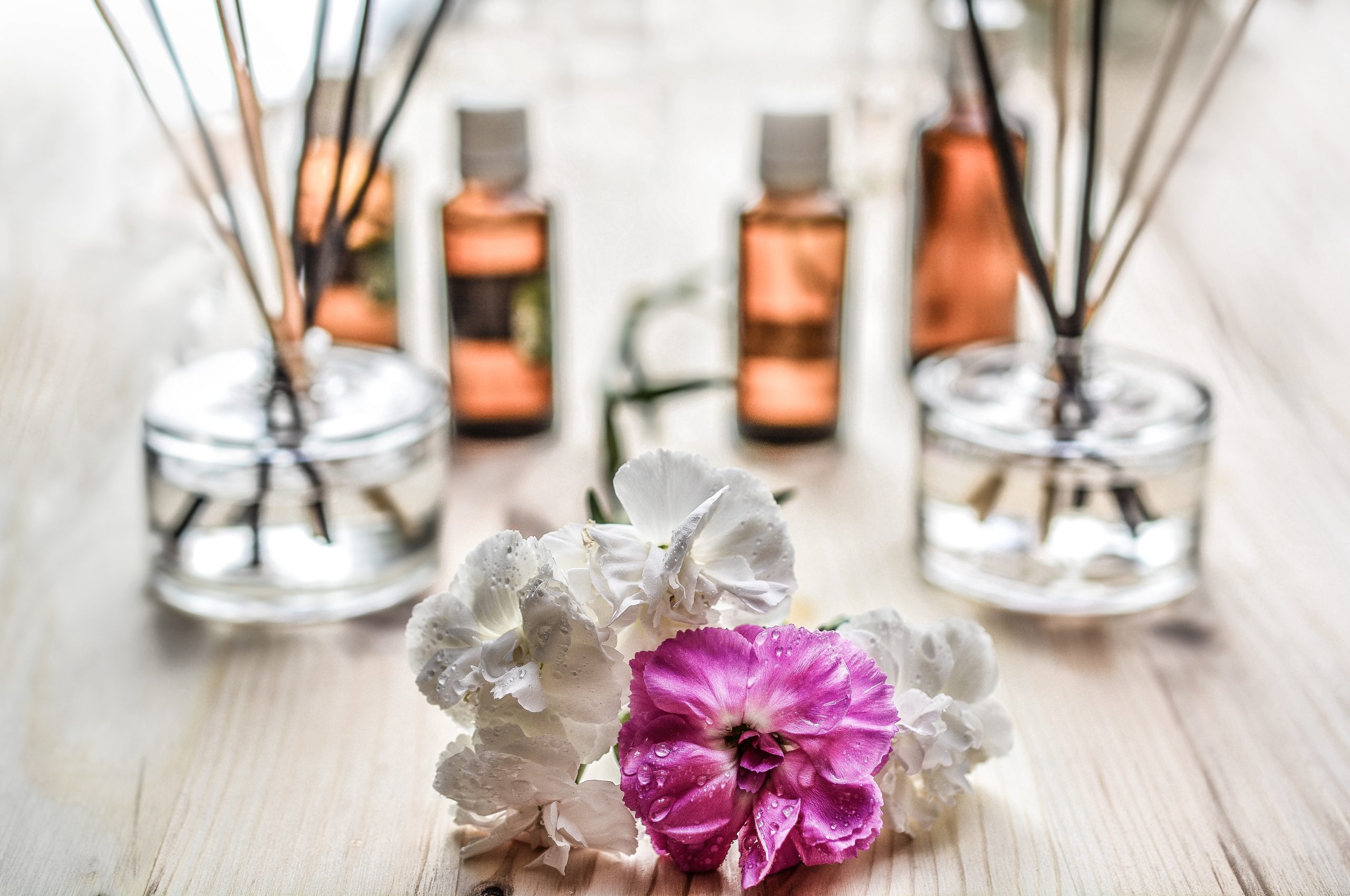 One purple and three white flowers on a light brown table with glass vases that have reed sticks in them and four bottles with essential oils