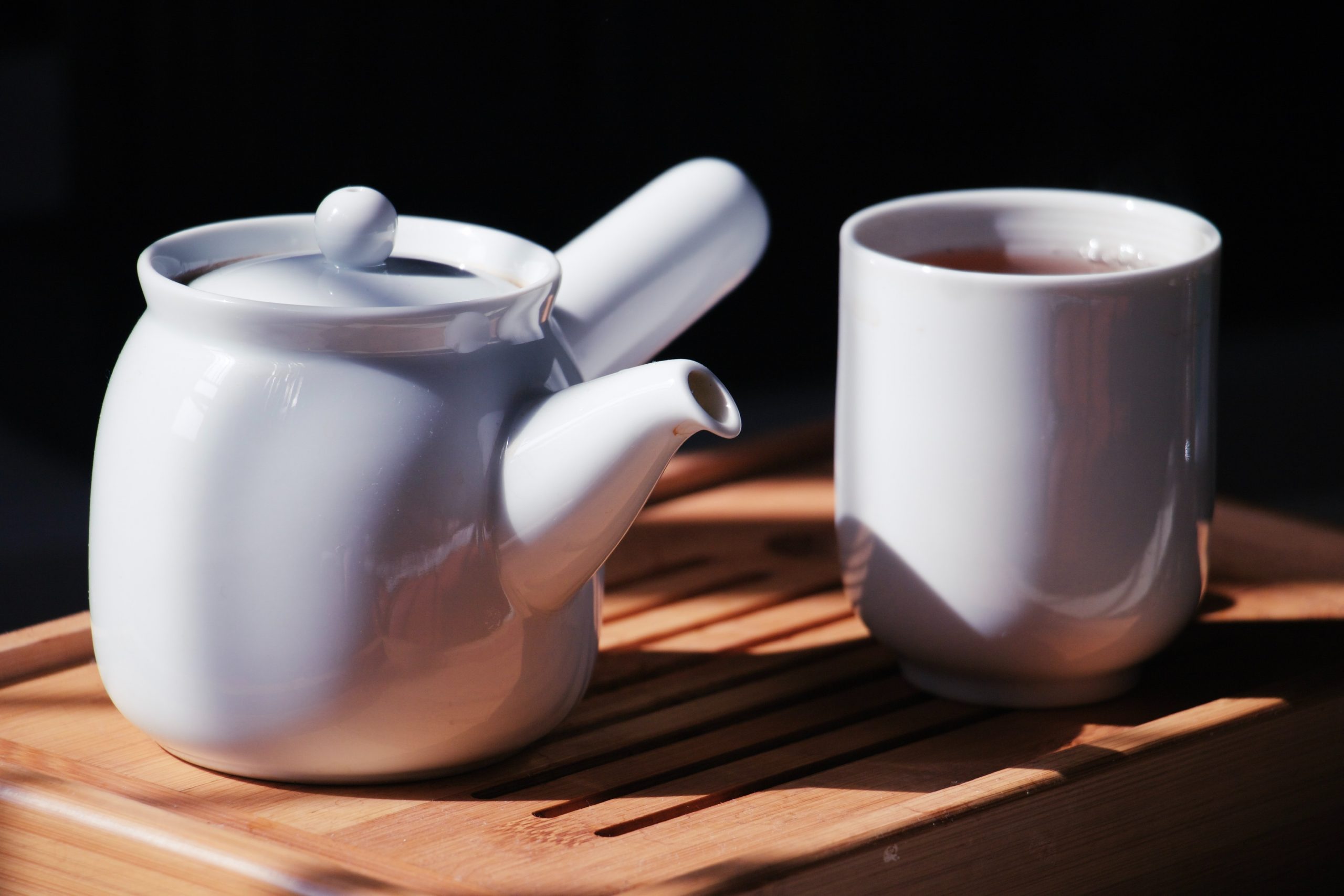 A porcelain teapot and cup filled with tea on a brown surface against a black background