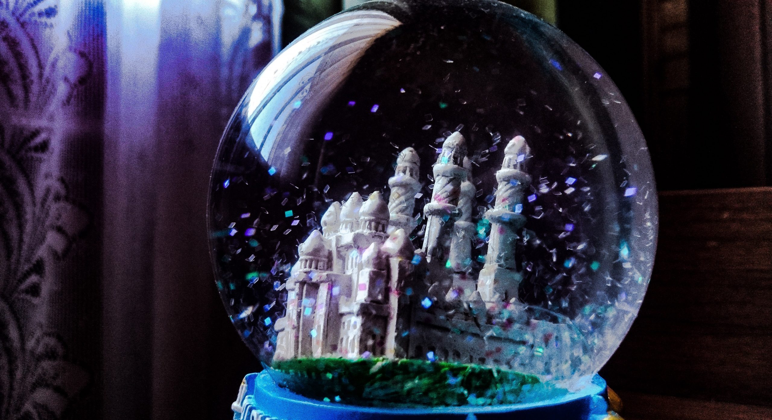 A snow globe with a castle and greenery inside it resting on a blue stand with purple curtains and a brown wooden cupboard behind it