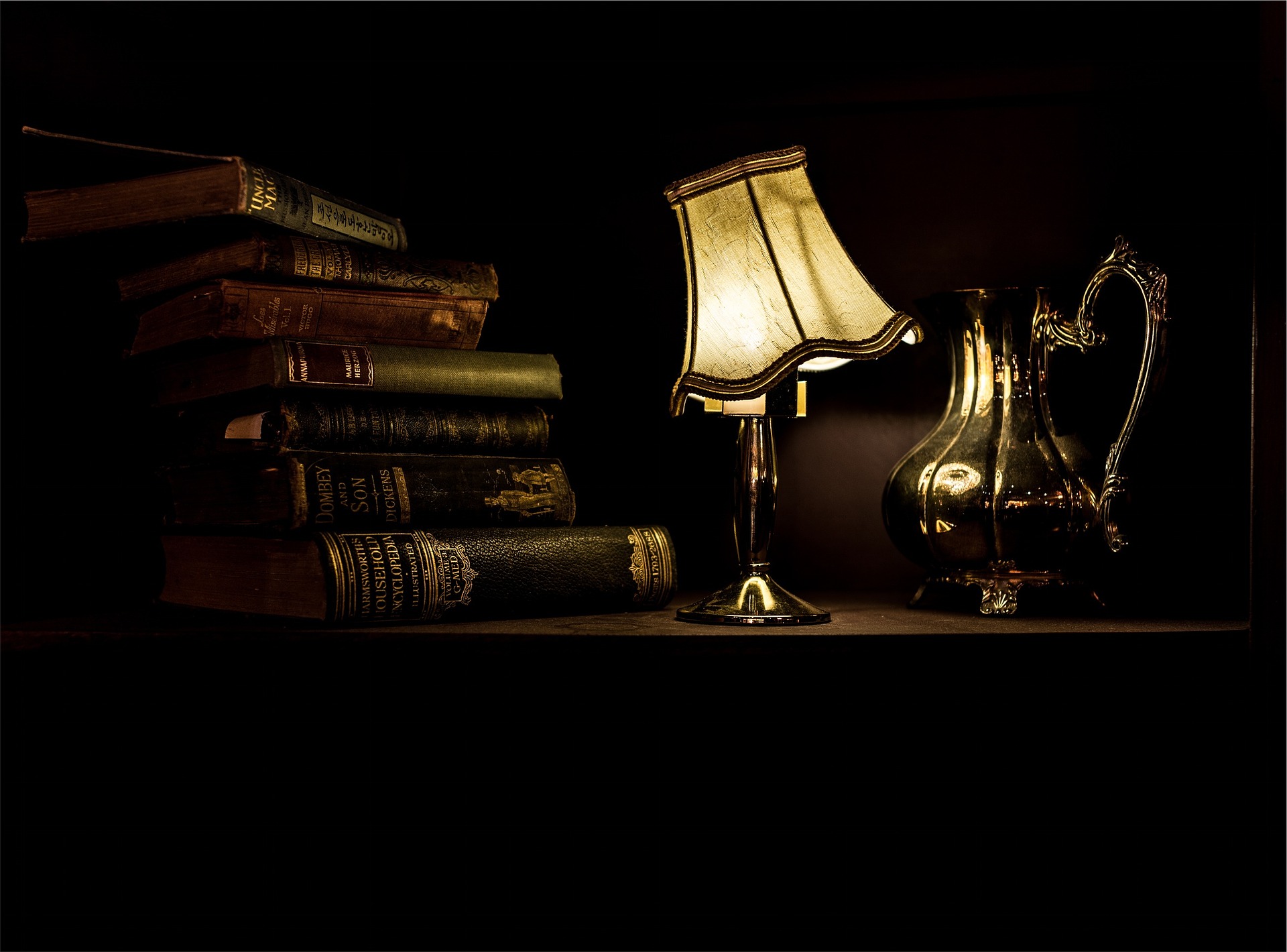 A lit desk lamp, hardcover books and a teapot on a desk.