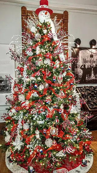 A Christmas tree adorned with a variety of red decorative elements and topped off with a snowman with a red nose and red hat.