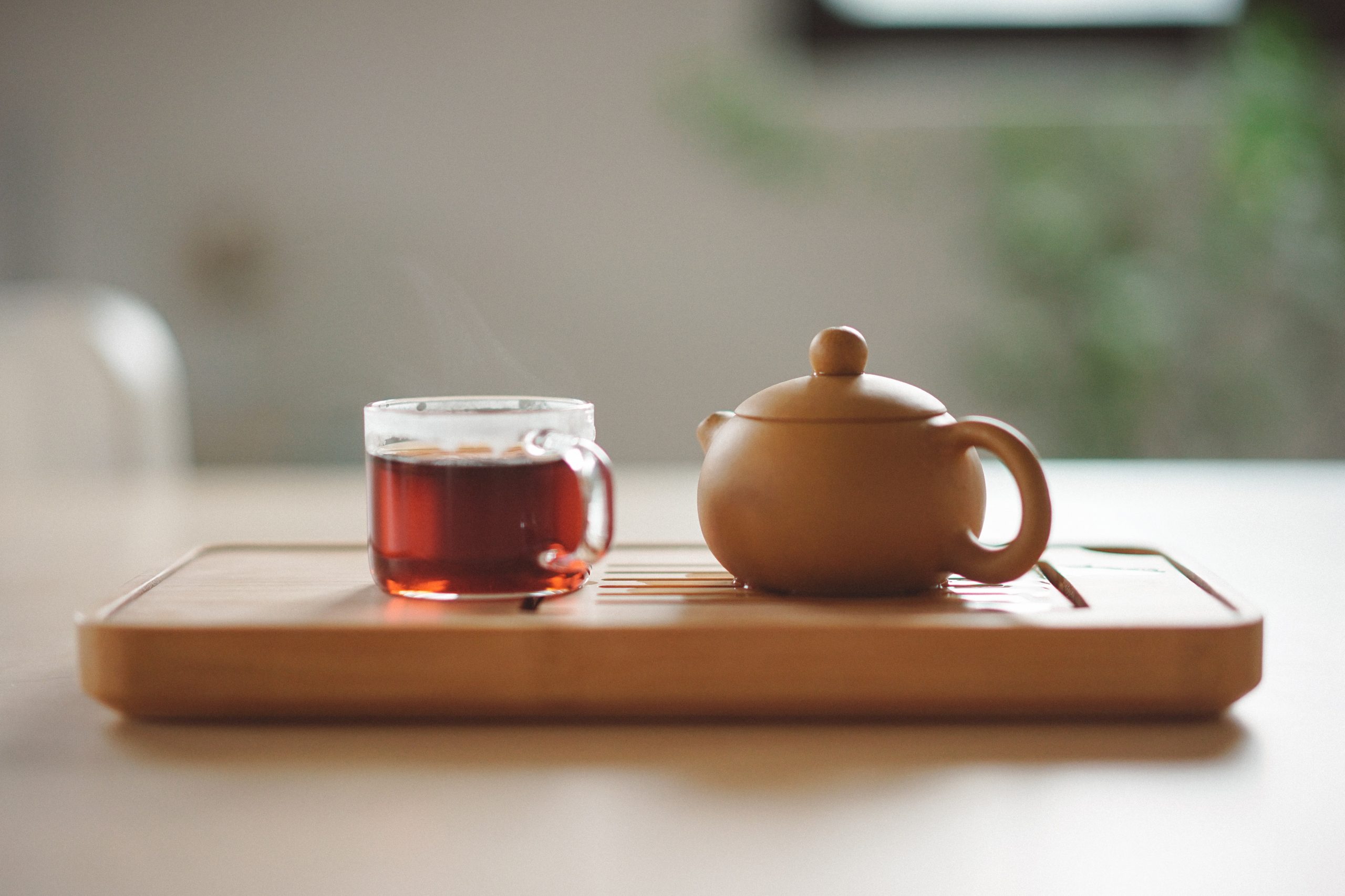 A cup of tea and a kettle placed side by side on a wooden tray, which is placed on a table.