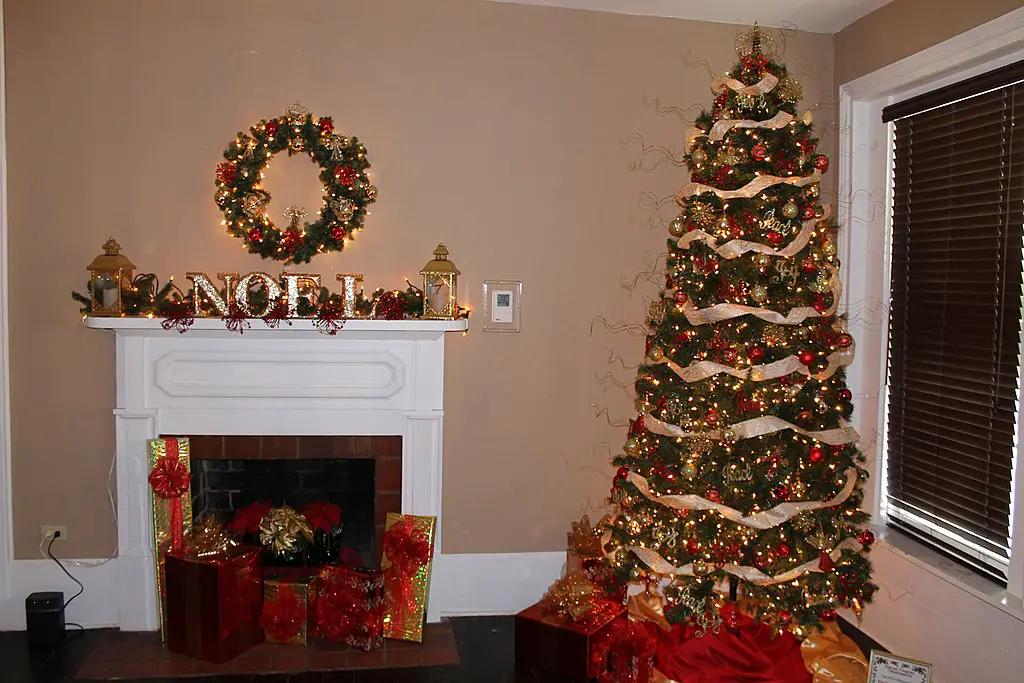 A Christmas tree placed in the corner of a room beside a decorated fireplace with numerous gifts.