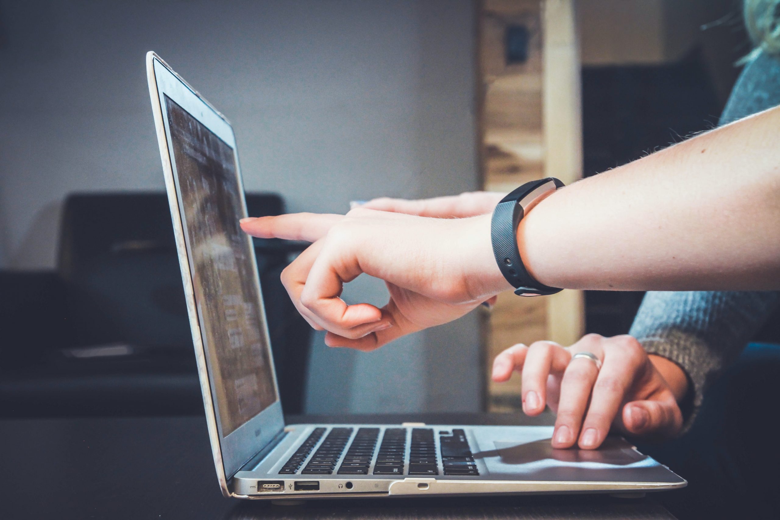 Two people using a laptop, with one of them wearing a fitness band who is also pointing a finger at the laptop screen.