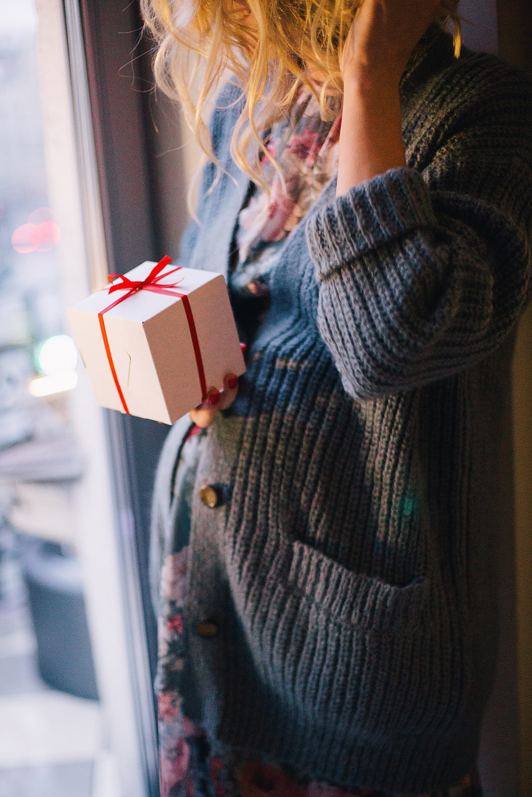 A pregnant woman dressed in a grey sweater holding a gift packed in a white box with a red ribbon