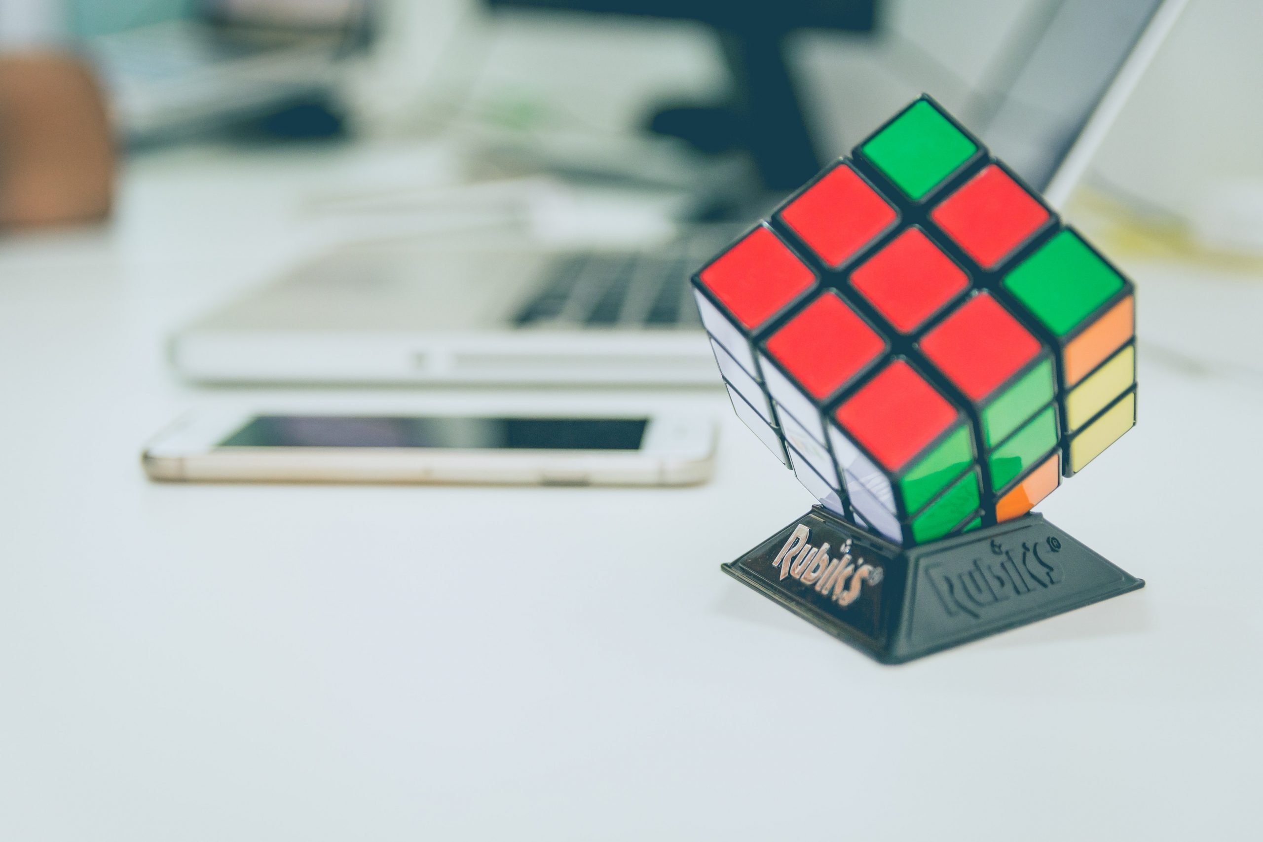 A Rubik’s cube in front of a smartphone and a laptop, with the bottom end of a black monitor also visible in the background.