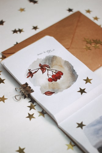 Clipped pages of a diary with a leaf and bunch of red berries drawn along with golden stars stuck on it set against a brownish gold envelope and white background with more golden stars on it