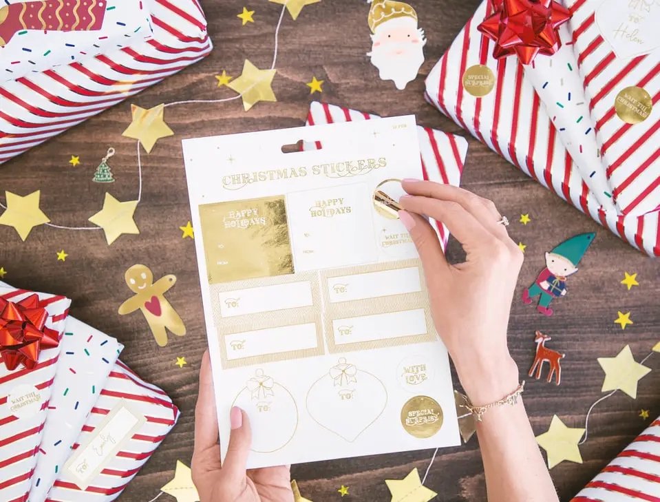 A person’s hand taking a gold sticker from a pad of gold stickers against a brown table background with golden star bunting and red and white gift wrapping