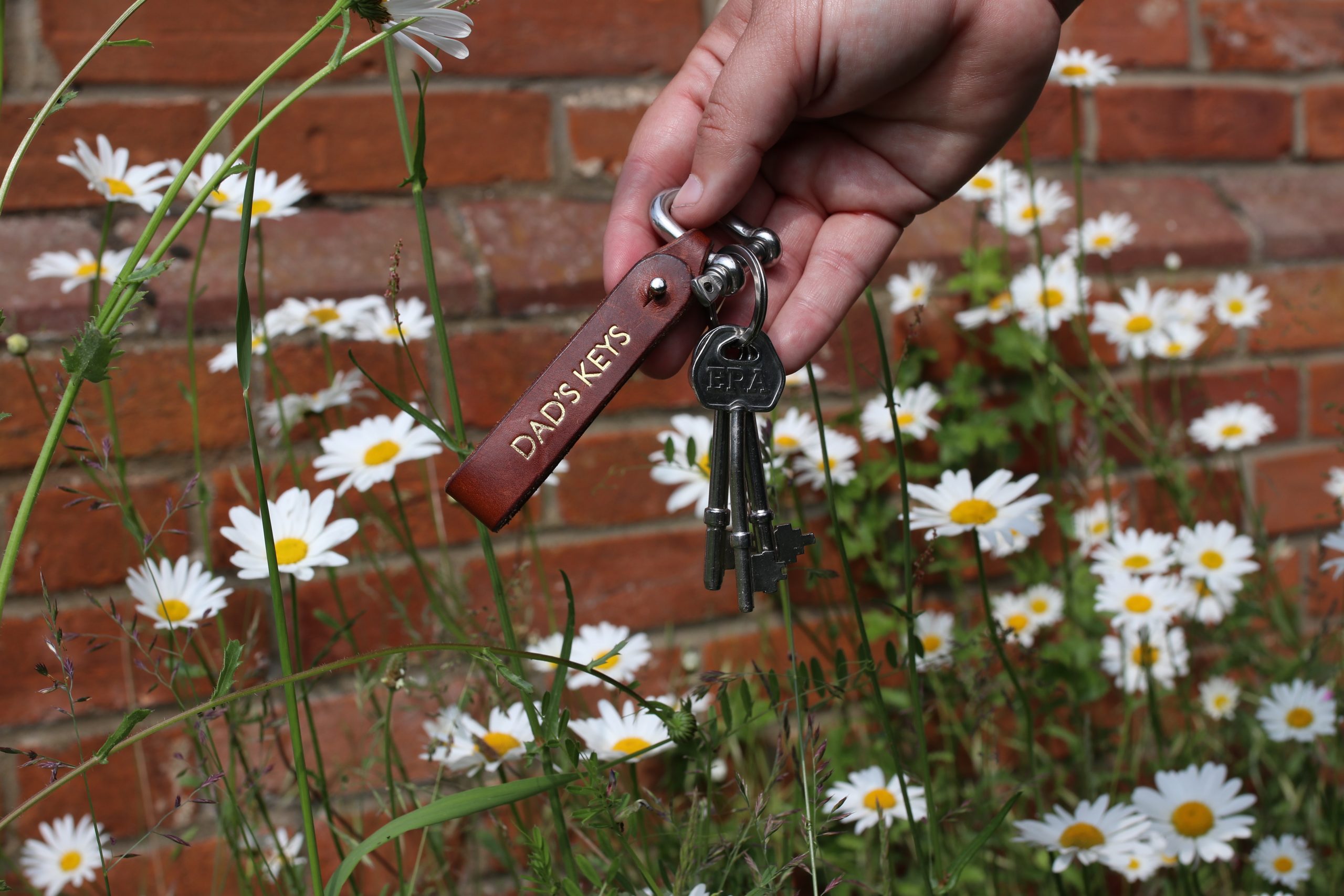 A hand holding a key chain that says DAD’S KEYS; flowers in the background.