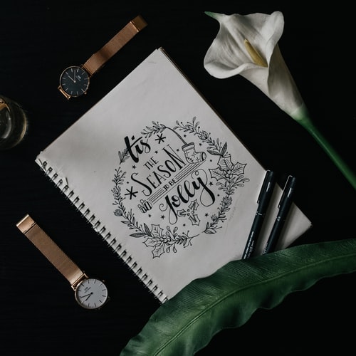 Page from a bullet journal An open journal page that reads, “Tis the season to be jolly” against a black background with two watches, two pens, and a flower.