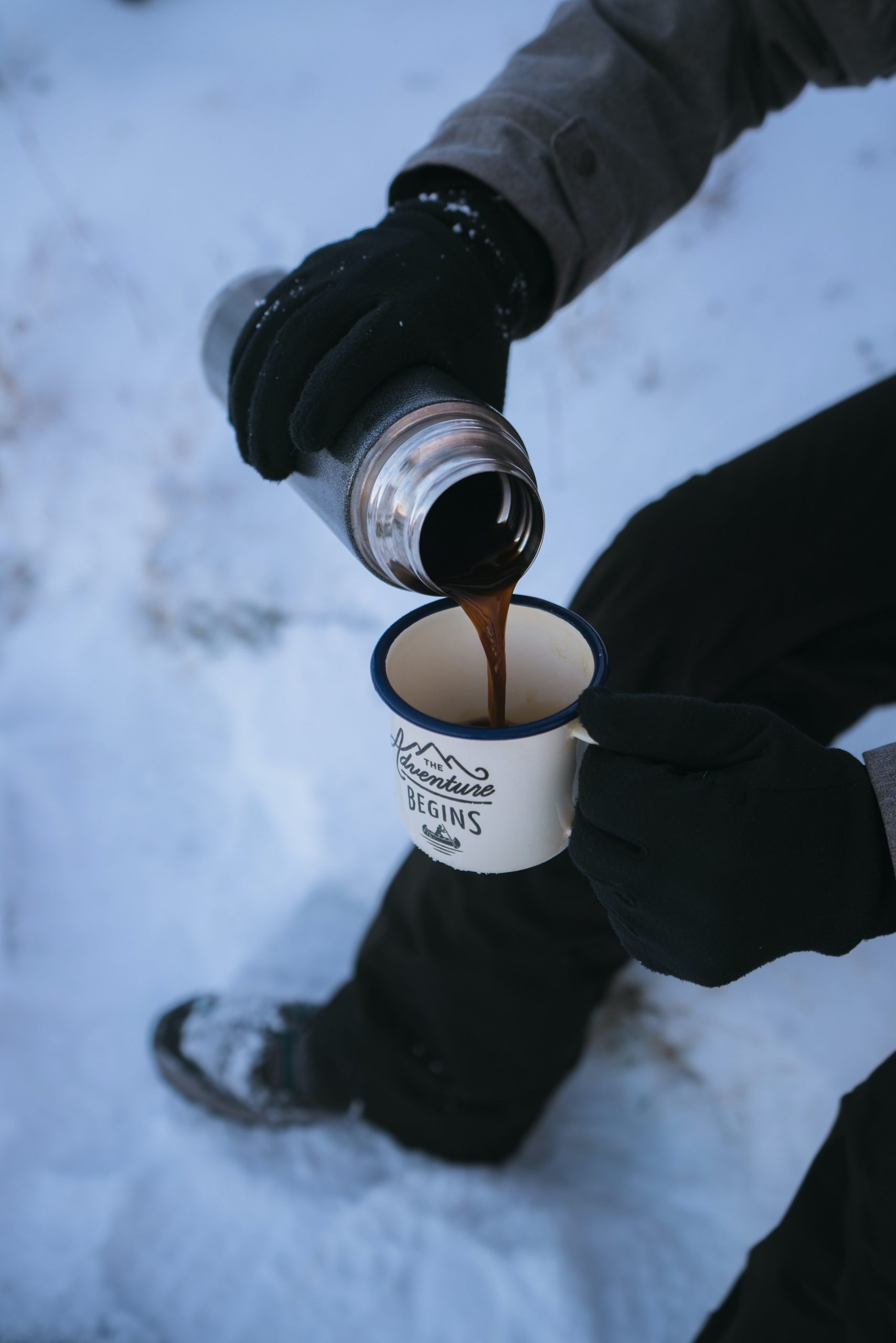 An individual wearing gloves pouring coffee into a coffee mug from an insulated bottle.