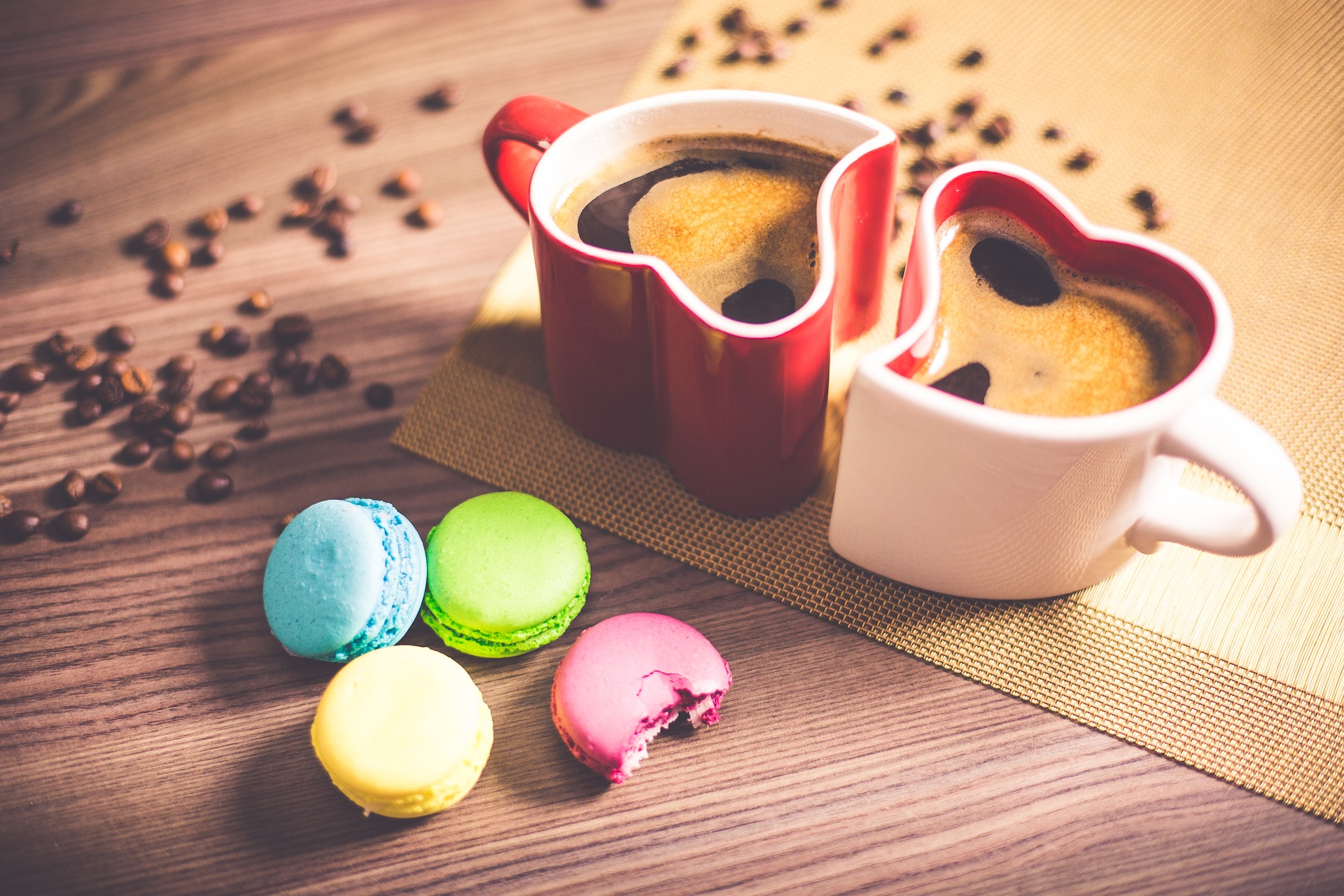 Two heart-shaped coffee mugs with hot coffee, macrons and coffee beans on a table.