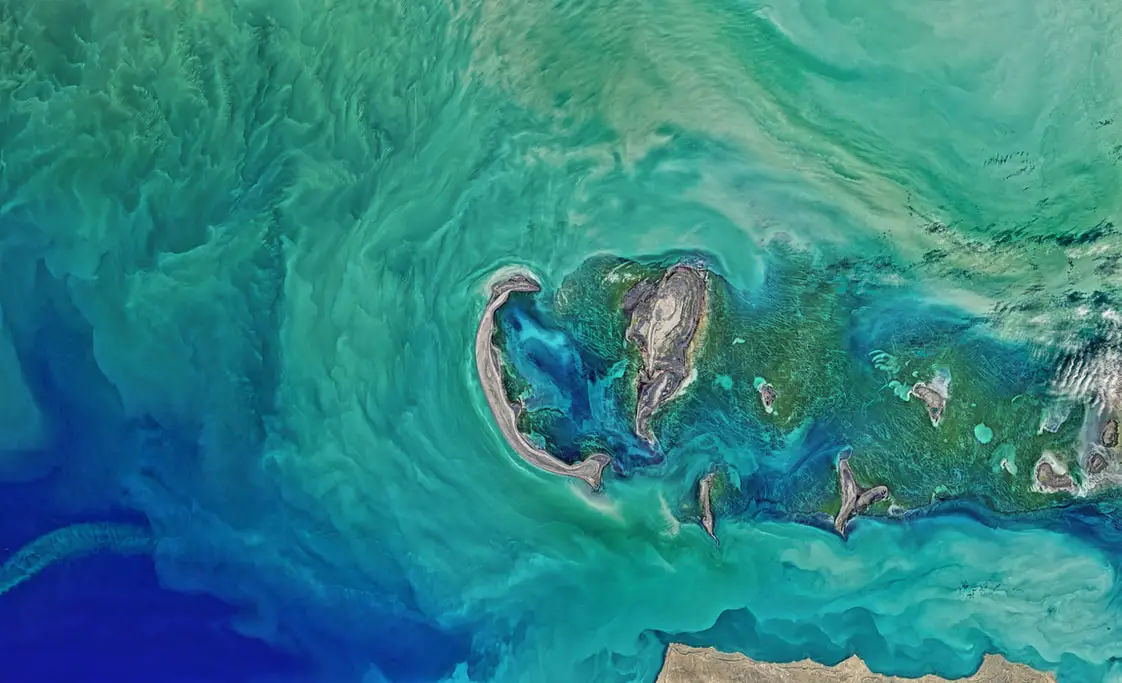 Aerial image of the ocean, showcasing spectacular blue shades all in one frame. In the center, you can see an island of sand.